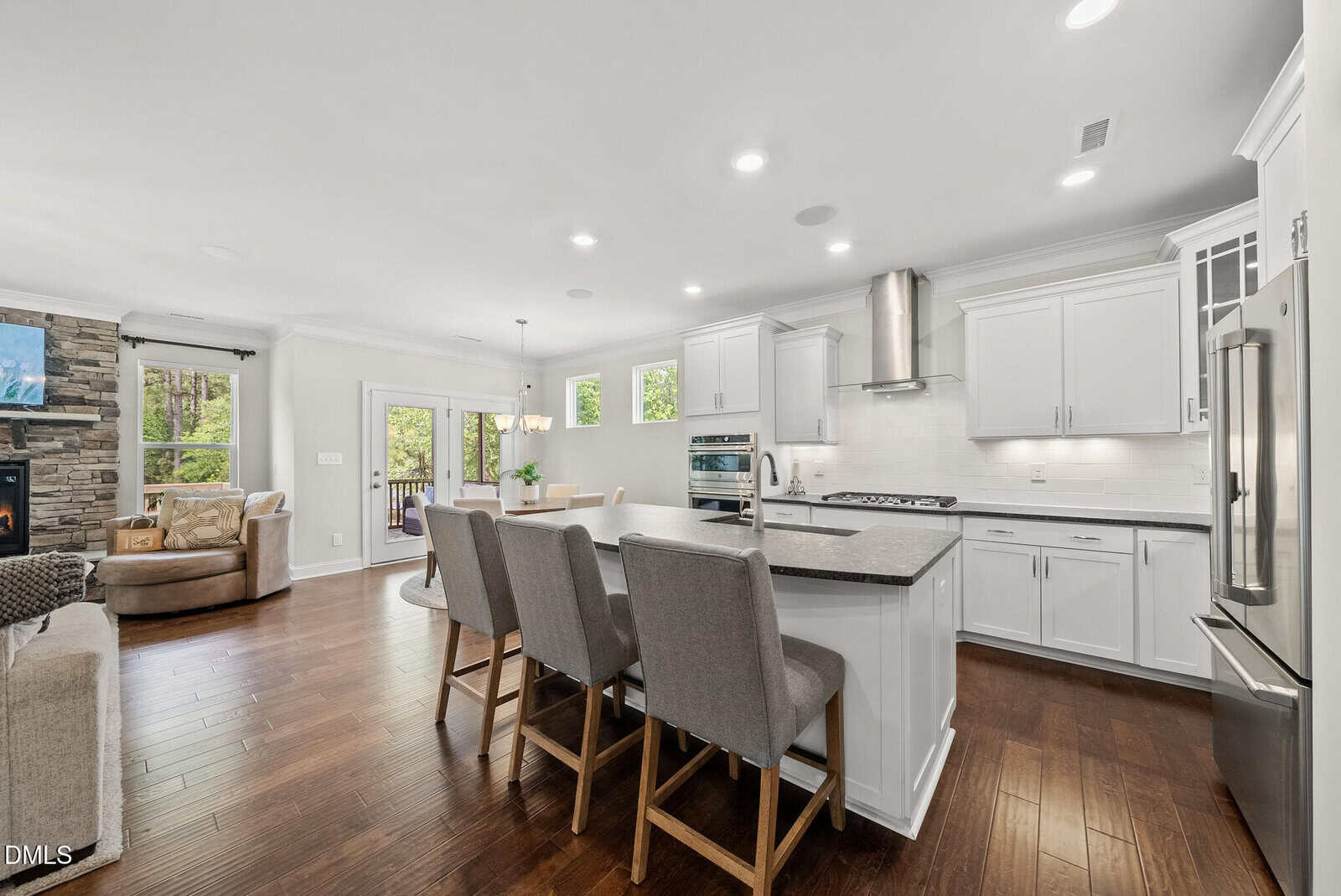 Modern open kitchen and living area with white cabinets, stainless steel appliances, island with gray chairs, wood floors, dining table, stone fireplace, and large windows showing trees outside.