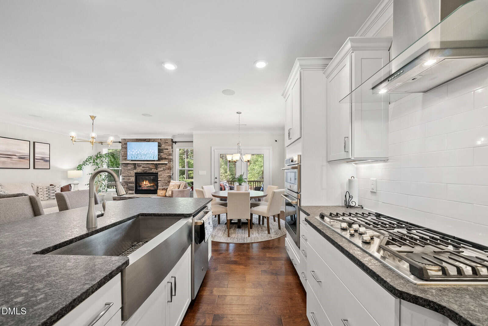 Modern kitchen with white cabinets, stainless steel appliances, farmhouse sink, and gas cooktop. Open layout leads to dining area and living room with fireplace, TV, and large windows offering natural light.