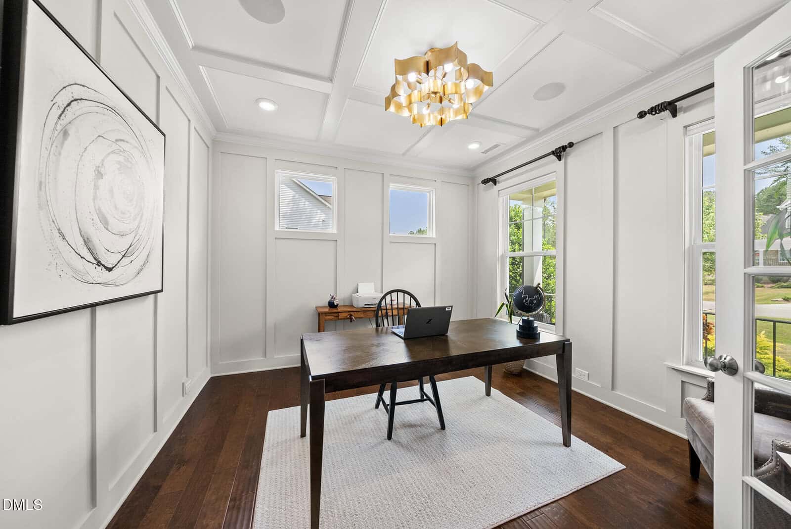 Bright home office with a wooden desk, laptop, chair, modern art on the wall, large windows, hardwood floor, and a unique gold chandelier.