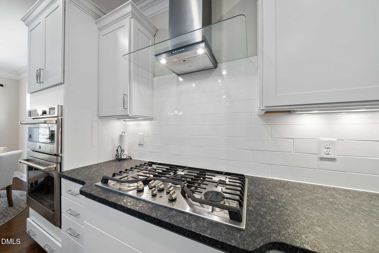 Modern kitchen with a black countertop, stainless steel gas stove, white cabinets, built-in oven, and a glass range hood against a white subway tile backsplash.