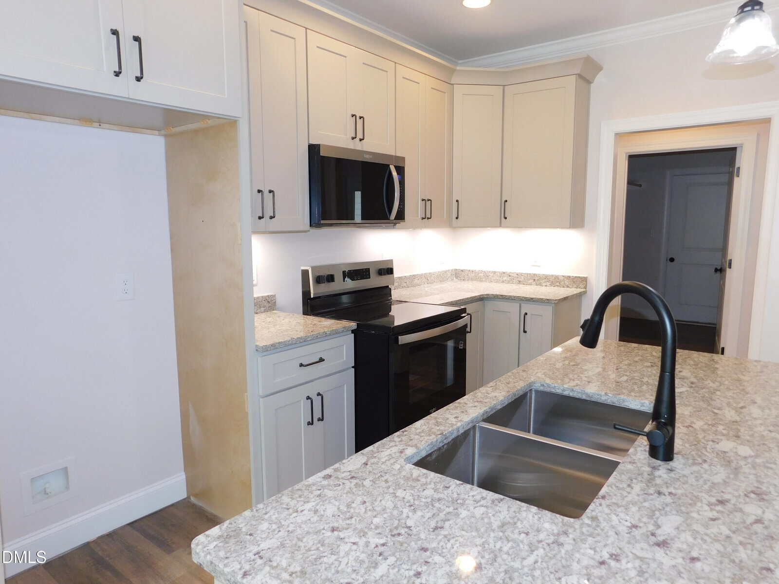 Modern kitchen with light cabinets, granite countertops, stainless steel appliances, a black faucet over a double sink, and an open space for a refrigerator on the left. Wood flooring and bright lighting are visible.
