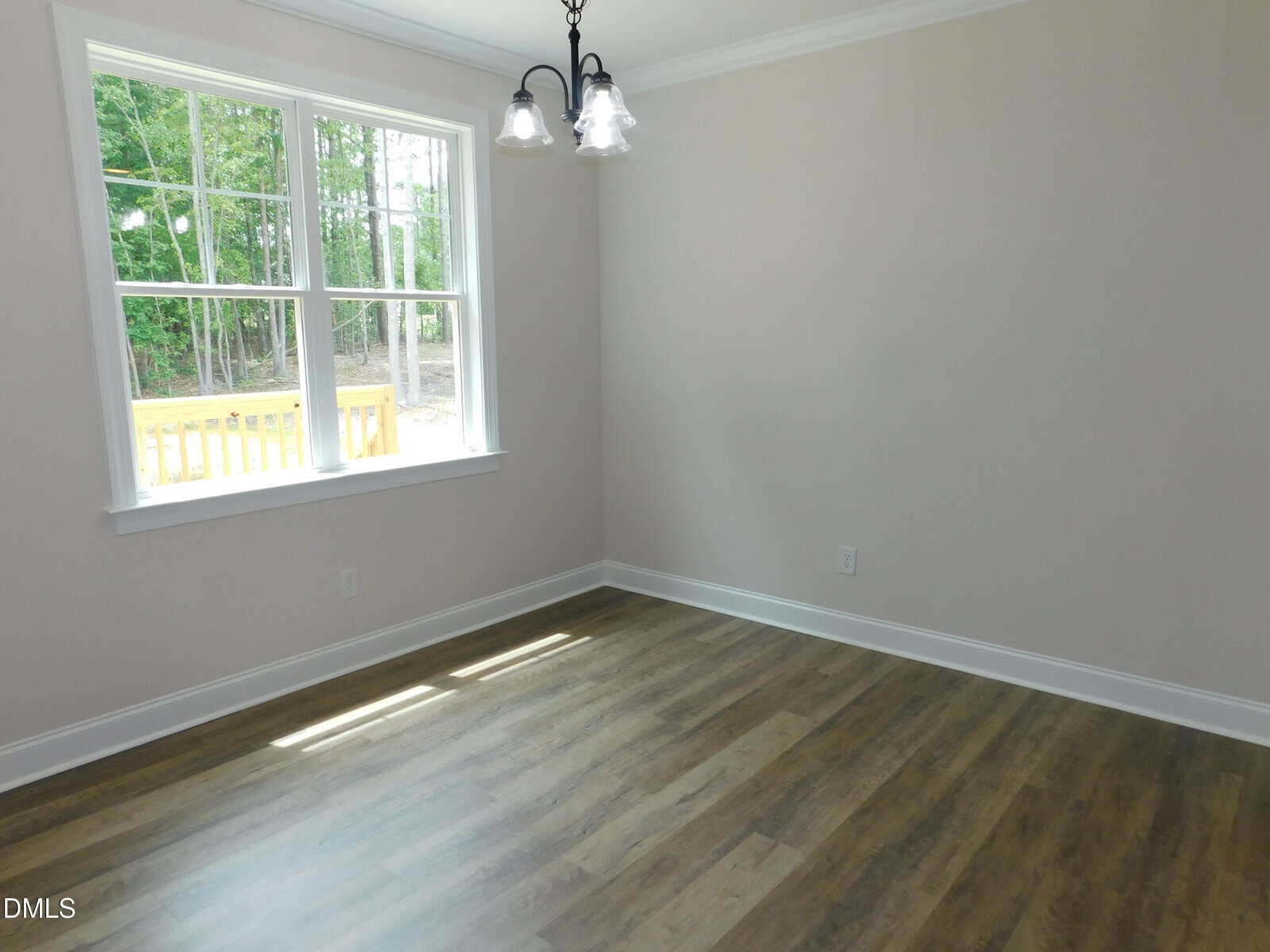 Bright, empty room with light beige walls, wood-look flooring, a three-light ceiling fixture, and a large window overlooking a deck and green trees outside.