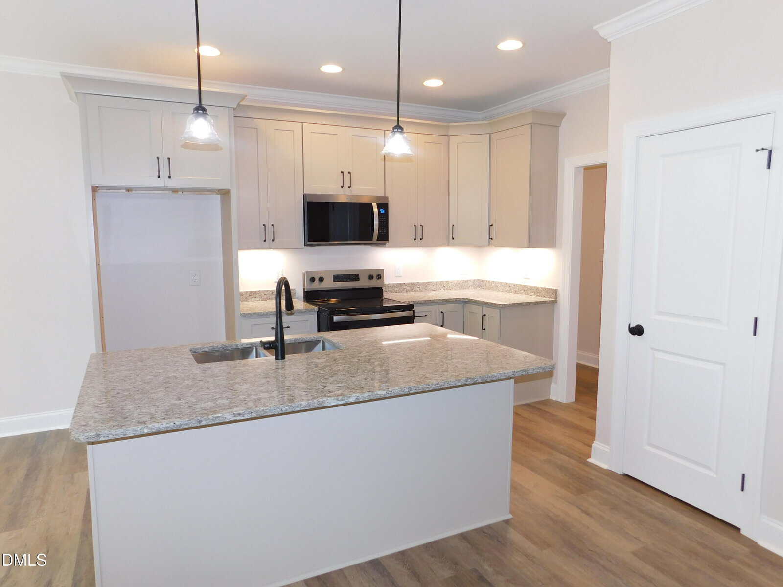 Modern kitchen with light gray cabinets, granite countertops, stainless steel appliances, an island with a sink, pendant lights, wood-style flooring, and white walls. A door and pantry are visible on the right side.