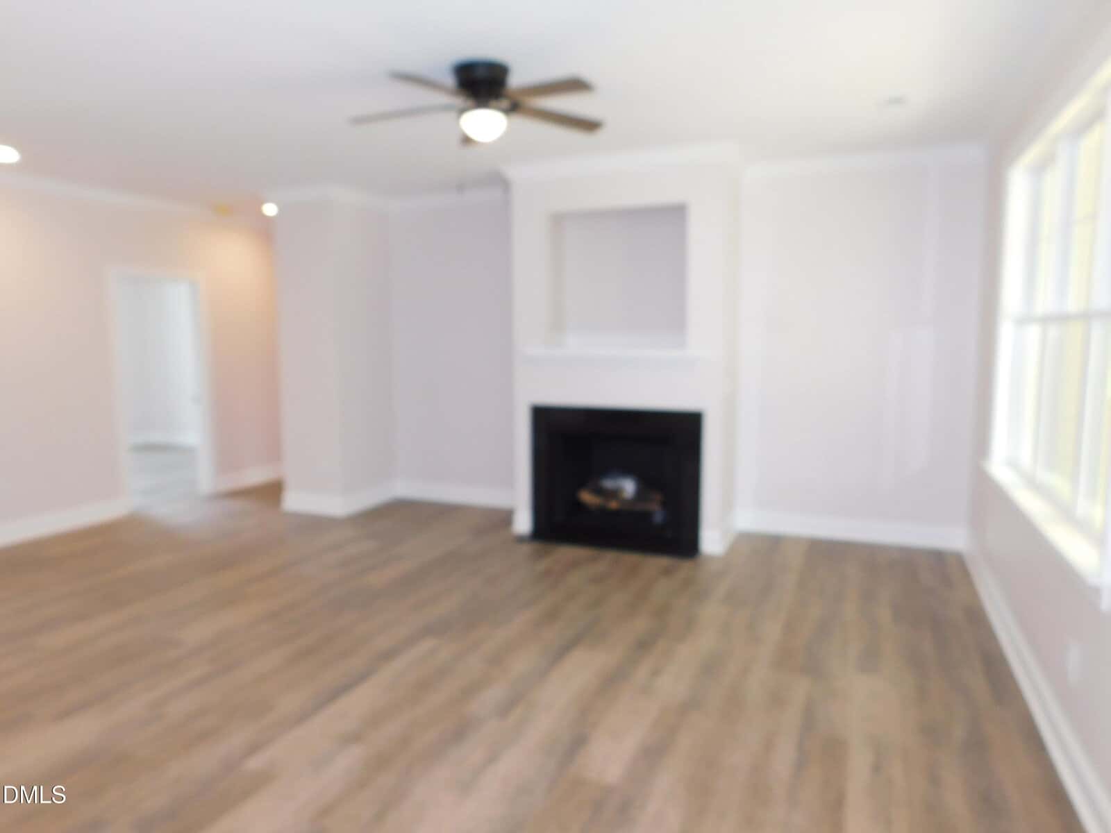 Blurry image of an empty living room with wood floors, a ceiling fan, a black fireplace, white walls, and a large window letting in natural light.