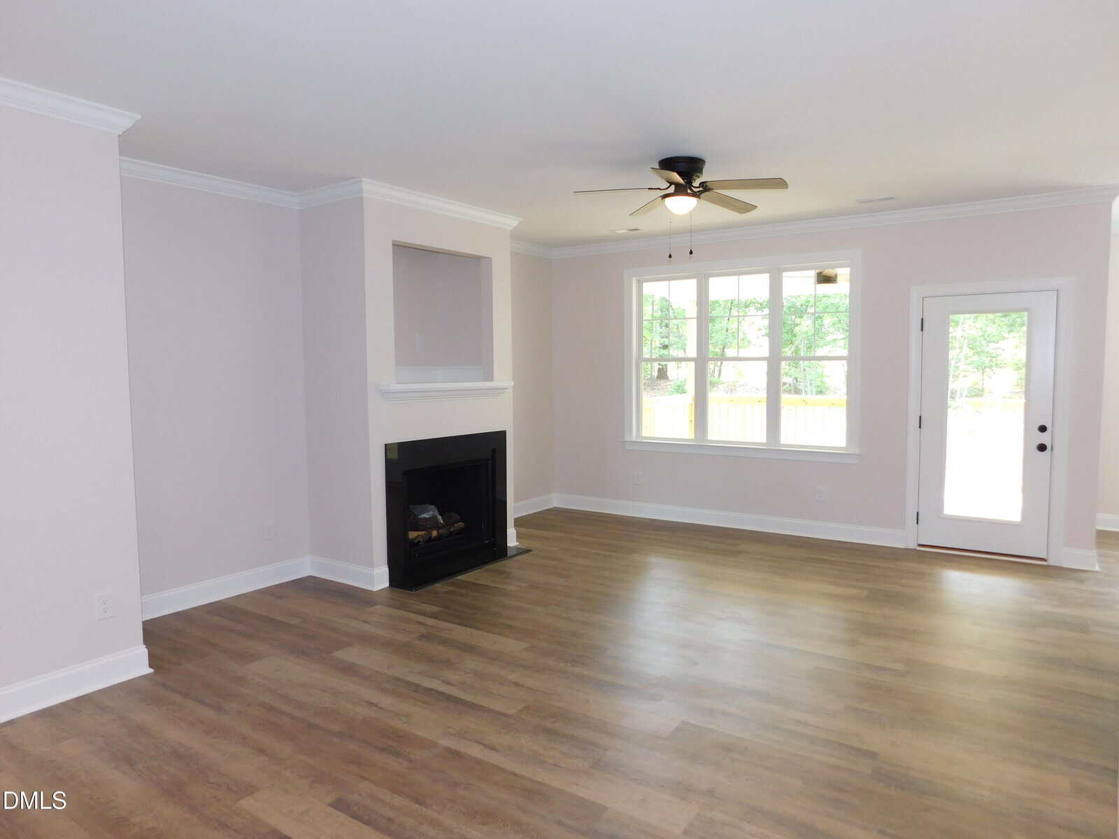 Bright living room with large window, ceiling fan, light walls, wood flooring, fireplace with mantel, and a door leading outside. The space is empty and well-lit by natural light.