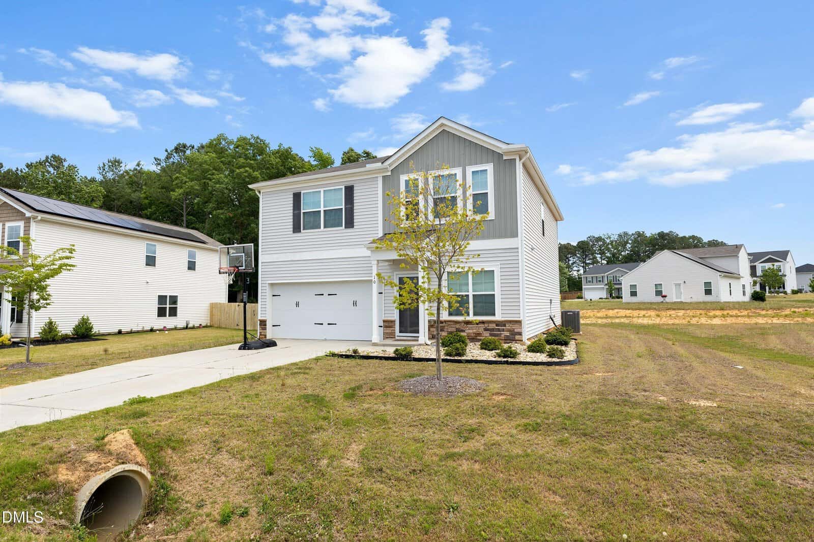 Two-story modern house with gray siding, white trim, and a garage. A small tree and landscaped plants are in the front yard. The house sits on a spacious lot with a concrete driveway and clear blue sky overhead.