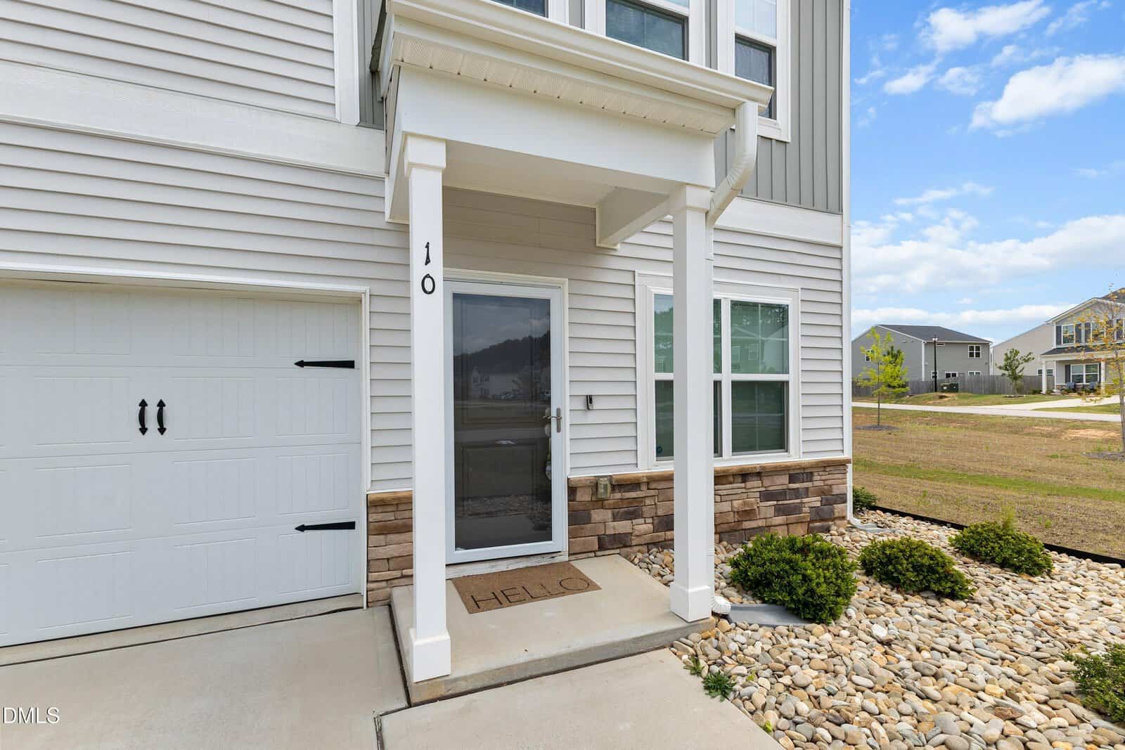 A modern house entryway with a small covered porch, white columns, and "HELLO" doormat. The exterior has light siding, stone accents, and is surrounded by landscaping with rocks and bushes. A garage door is on the left.