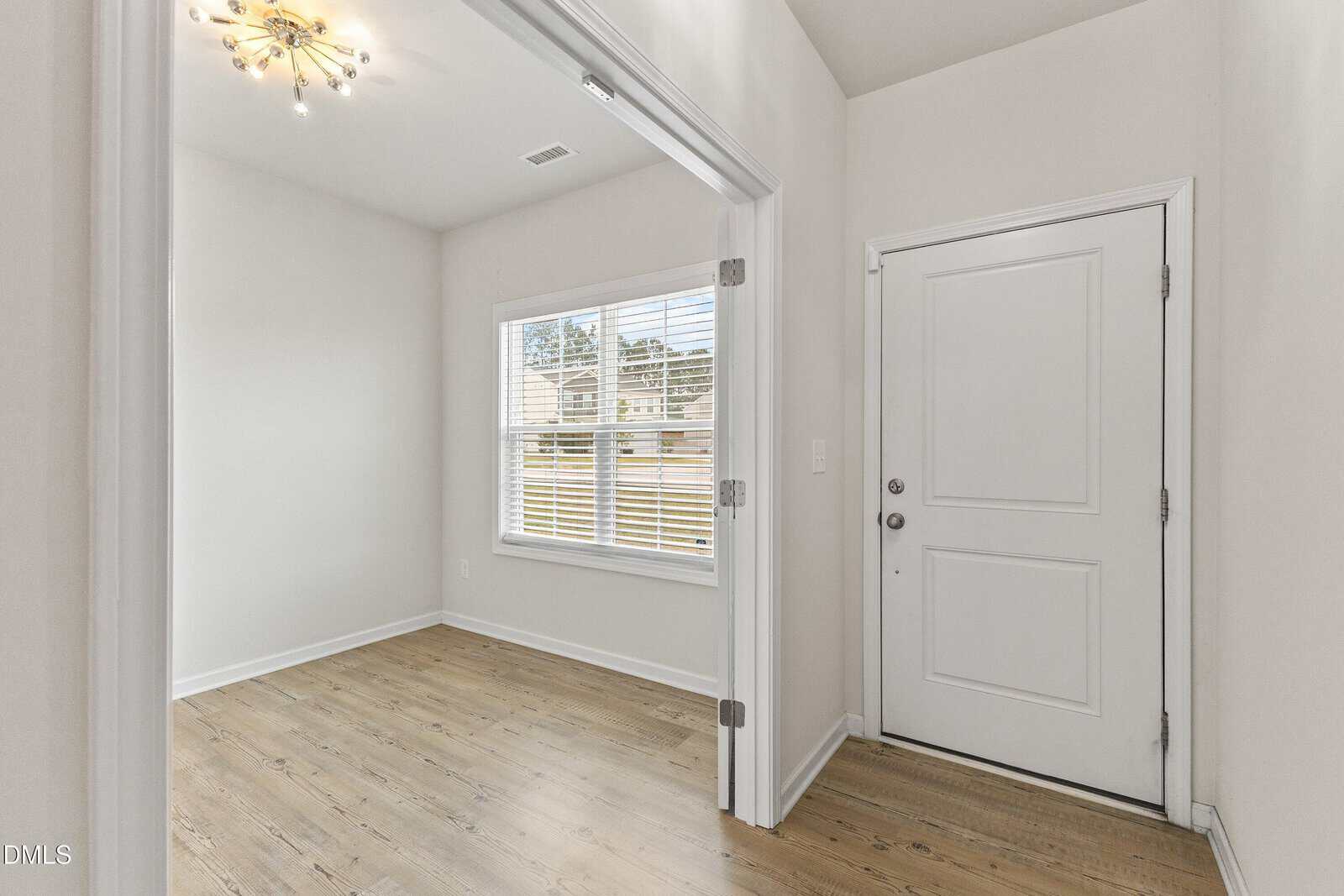 Bright entryway with wood flooring, a white front door, and an adjacent small room with glass double doors, a window with blinds, and a modern light fixture. Walls are painted light beige.