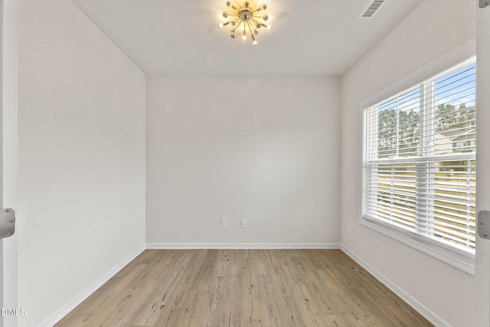 Bright, empty room with white walls, a modern chandelier on the ceiling, wood flooring, and a large window with blinds letting in natural light.