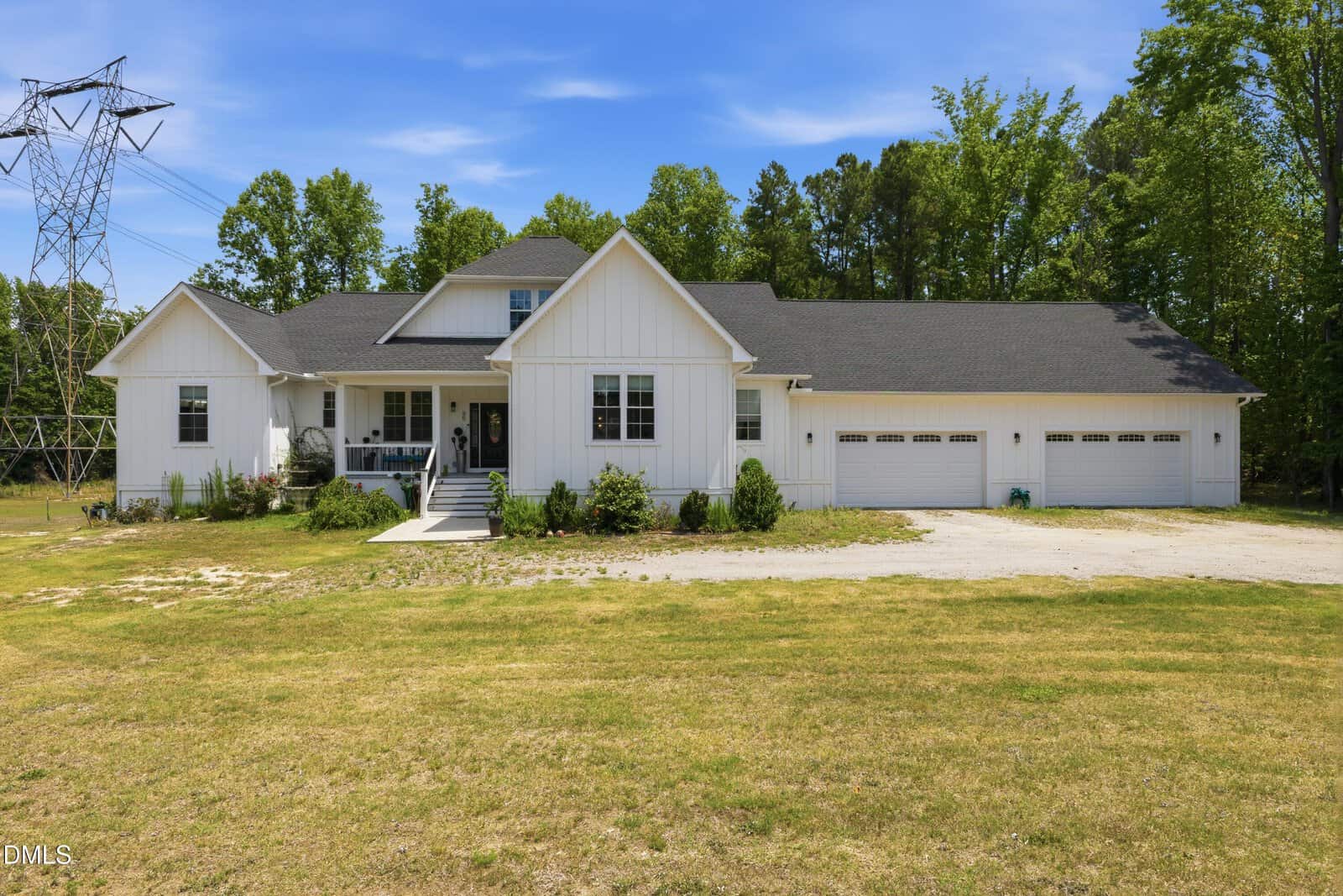 A modern white farmhouse on 65 Soggy Bottom Trail in Zebulon, featuring a large front porch, black roof, three-car garage, and landscaped yard, set against a backdrop of trees and a blue sky.