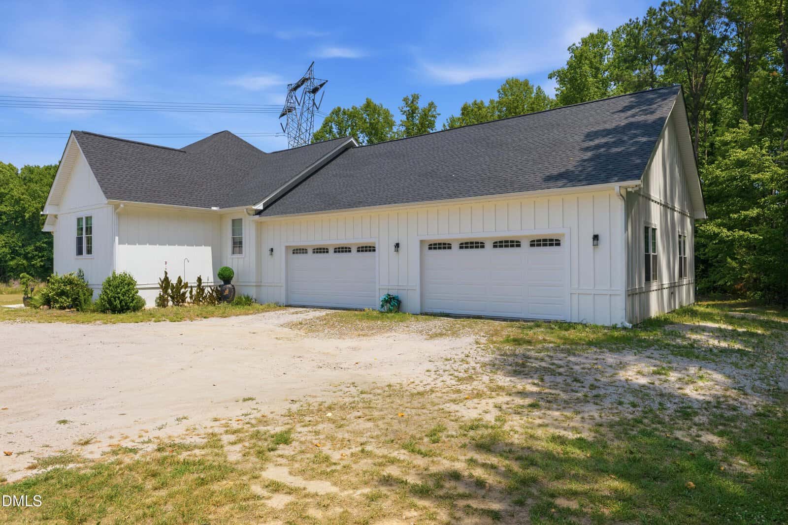 A white, single-story house with a dark roof and four-car garage sits by a gravel driveway, surrounded by trees and greenery on a sunny day.