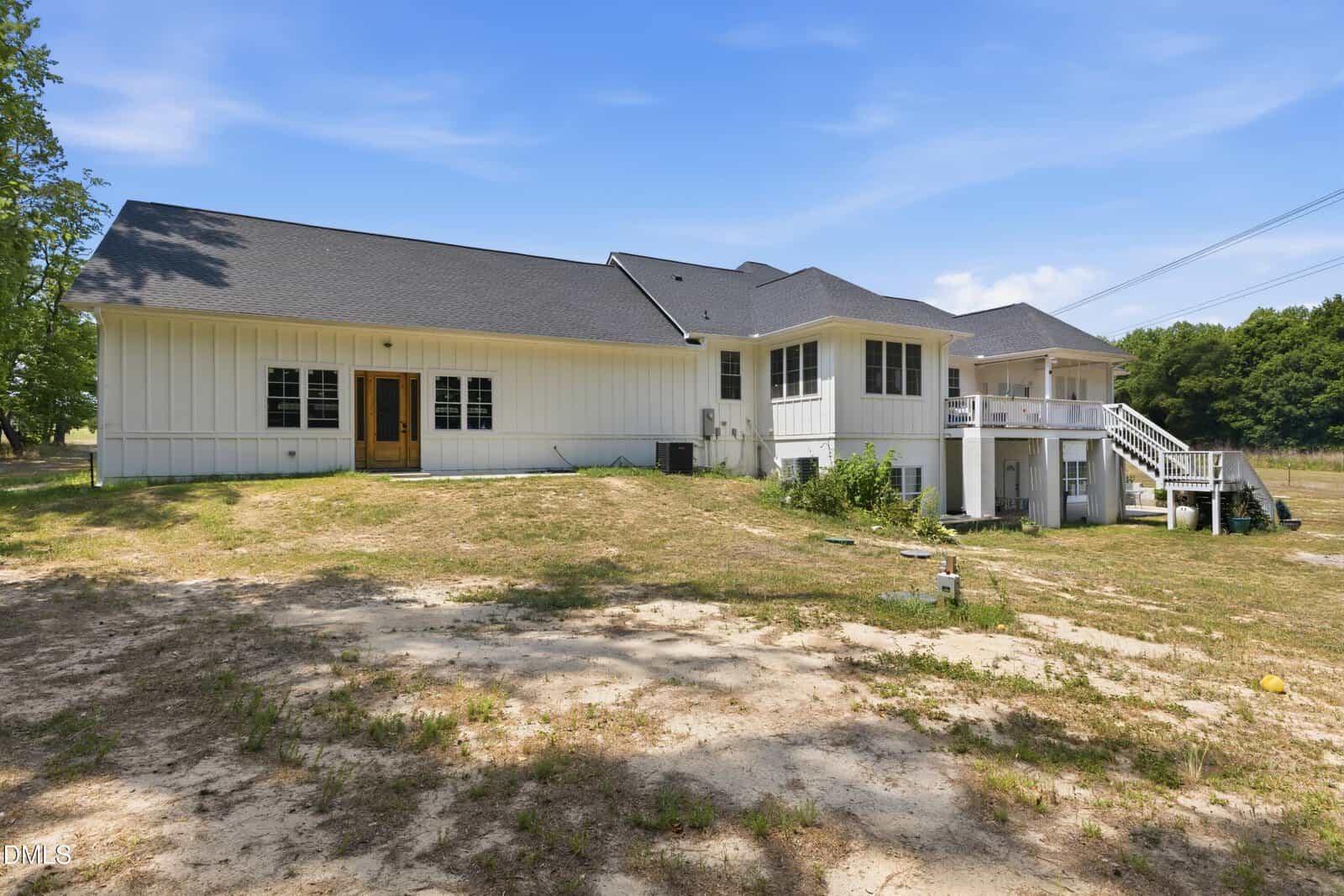 Single-story white house with dark roof, wooden French doors, screened porch, and stairs leading to a raised deck. The yard is sandy with patches of grass, surrounded by trees under a clear blue sky.