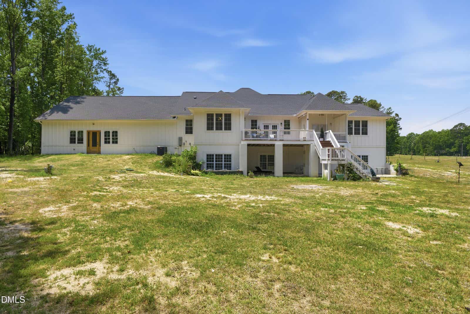 A large white house with multiple levels, a gray roof, and outdoor stairs leading to a covered patio area, surrounded by grassy open land and trees under a blue sky.