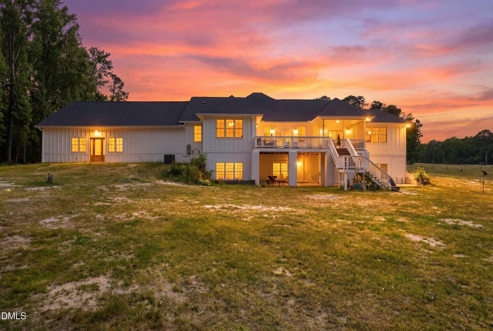 A large white house with illuminated windows sits on a grassy field at sunset, with an expansive deck, stairs, and trees in the background under a colorful sky.
