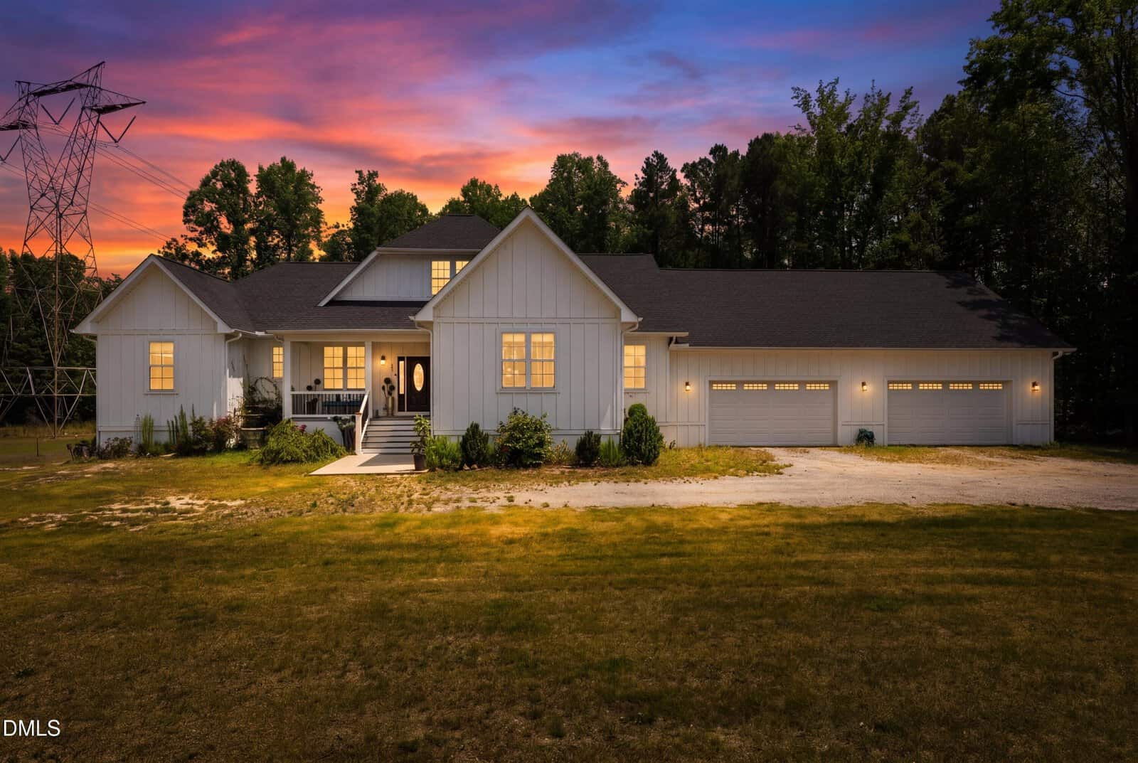 A modern white house with warm lights glows at dusk, surrounded by trees and a grassy yard. The sky is vibrant with orange, purple, and blue hues, and a row of garage doors is visible on the right side.