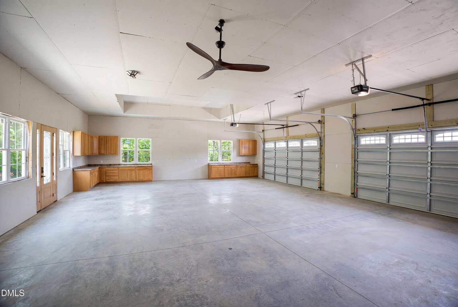 Spacious, clean three-car garage with light gray concrete floor, two ceiling fans, overhead garage doors, large windows, and wooden cabinets along the back wall. Natural light fills the room.