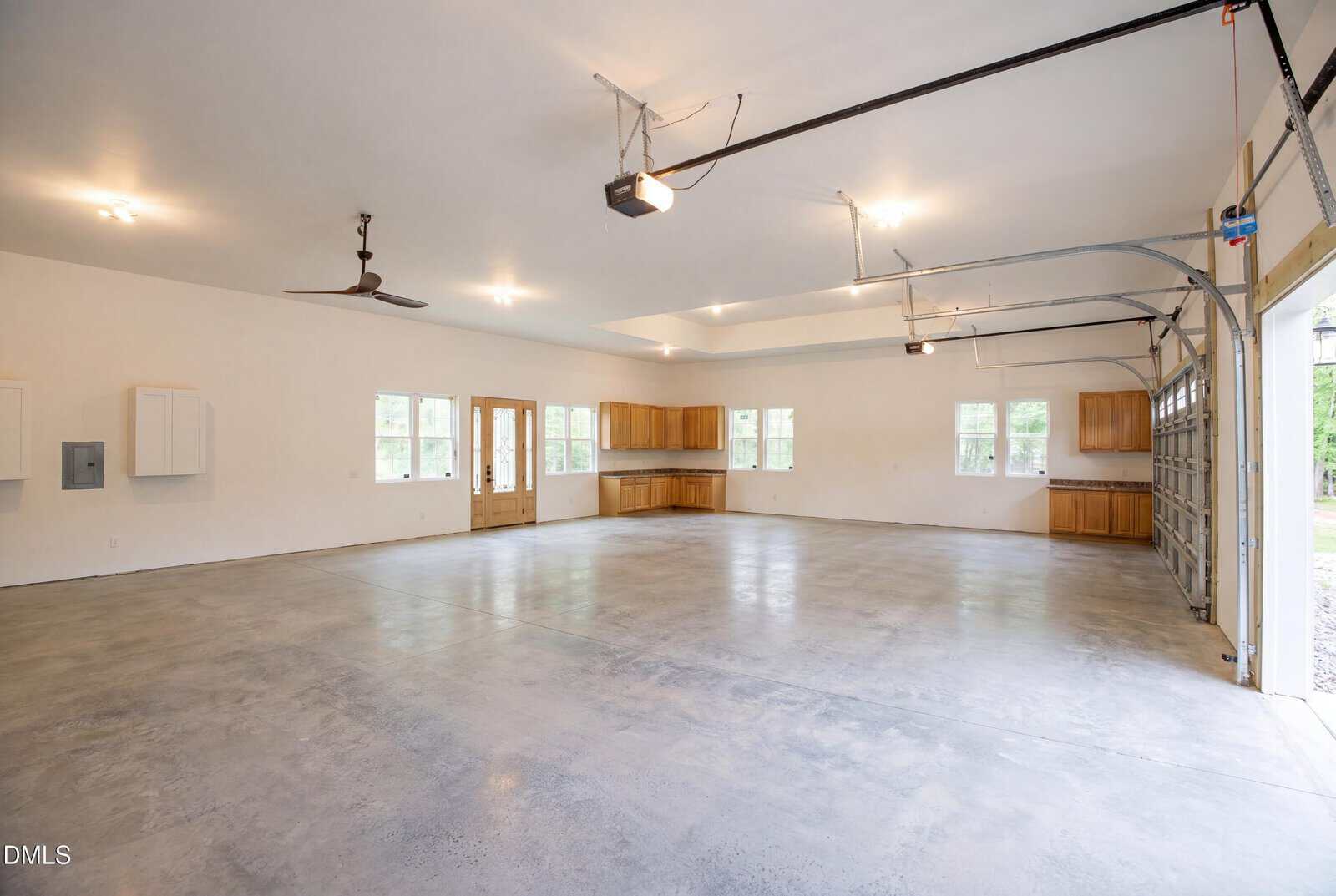 Spacious, empty garage with polished concrete floors, ceiling lights, wooden cabinets, windows, a ceiling fan, and open garage doors letting in natural light.
