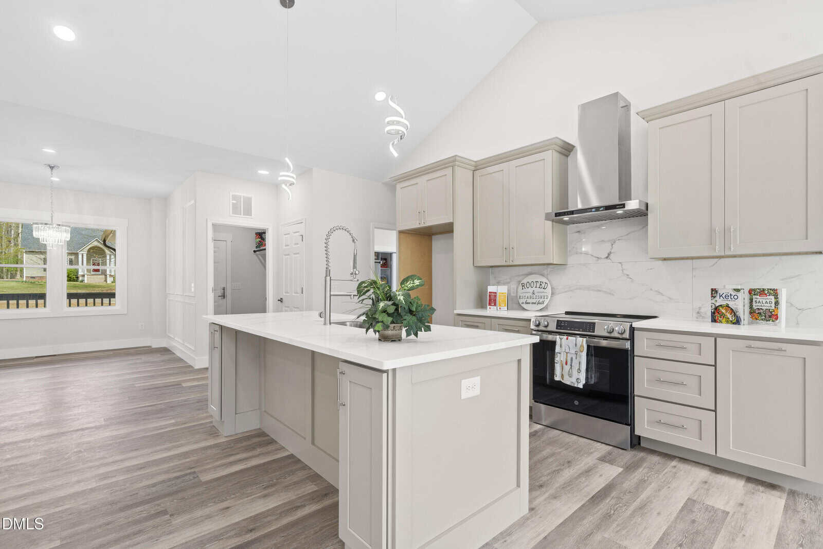 Modern kitchen with light gray cabinets, marble backsplash, stainless steel appliances, and a large island with a built-in sink and potted plant. Dining area and window with outdoor view are visible in the background.