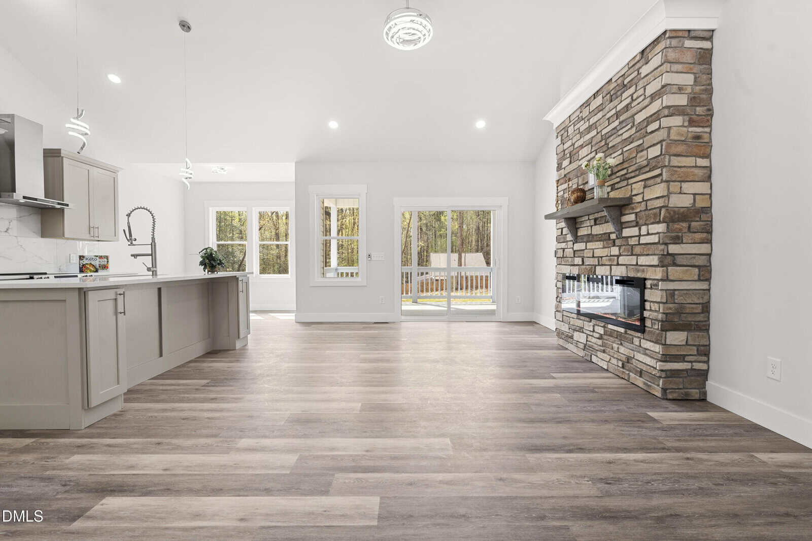 Modern open-concept living area with light wood floors, a stone fireplace, a spacious kitchen with gray cabinets, and large windows, leading to a backyard view.