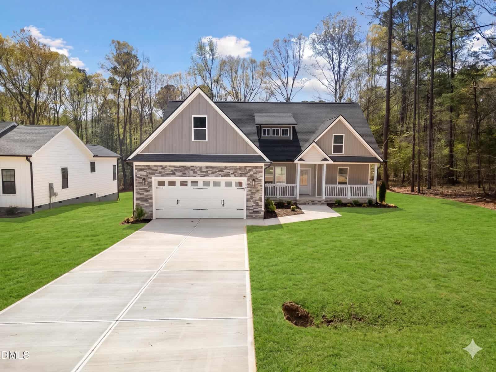 A modern two-story house with a gray exterior, stone accents, a front porch, and an attached two-car garage, this beautiful property at 116 Blackfoot Drive in Louisburg sits on a large green lawn with a concrete driveway and is surrounded by trees.