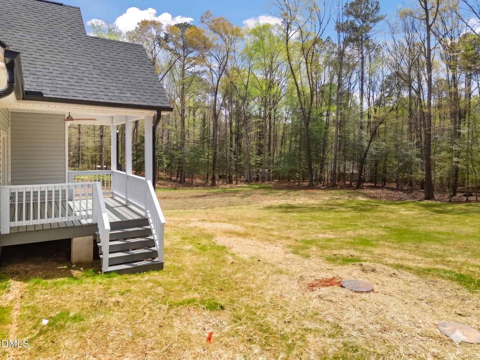 A white house with gray steps leads to a grassy backyard bordered by a forest. The yard has patches of bare ground, and a few utility covers are visible. Trees in the background have early spring foliage.