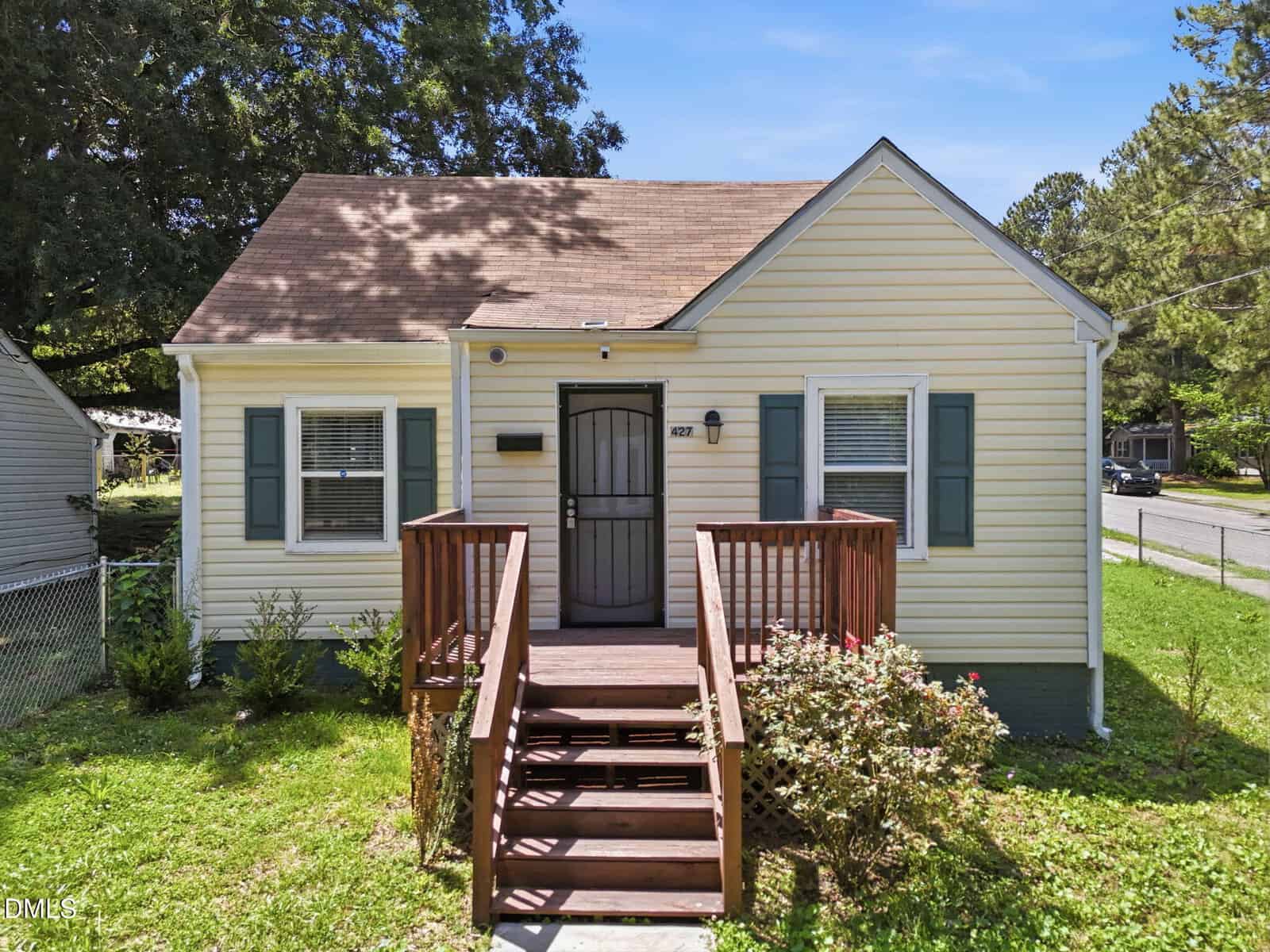 A small, light yellow house at 427 N Clark Street in Henderson, with green shutters and a brown shingle roof, features a wooden front porch with steps and metal door, surrounded by grass, shrubs, and trees on a sunny day.