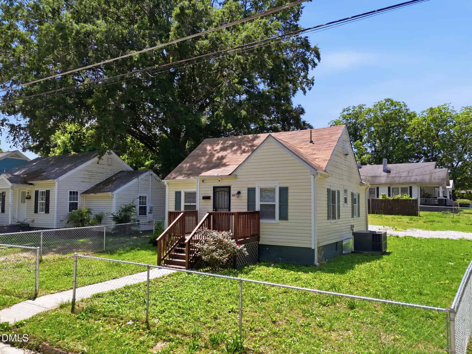 A small, single-story beige house with a brown roof and wooden front steps, surrounded by a chain-link fence and green lawn, with neighboring houses and tall trees in the background.