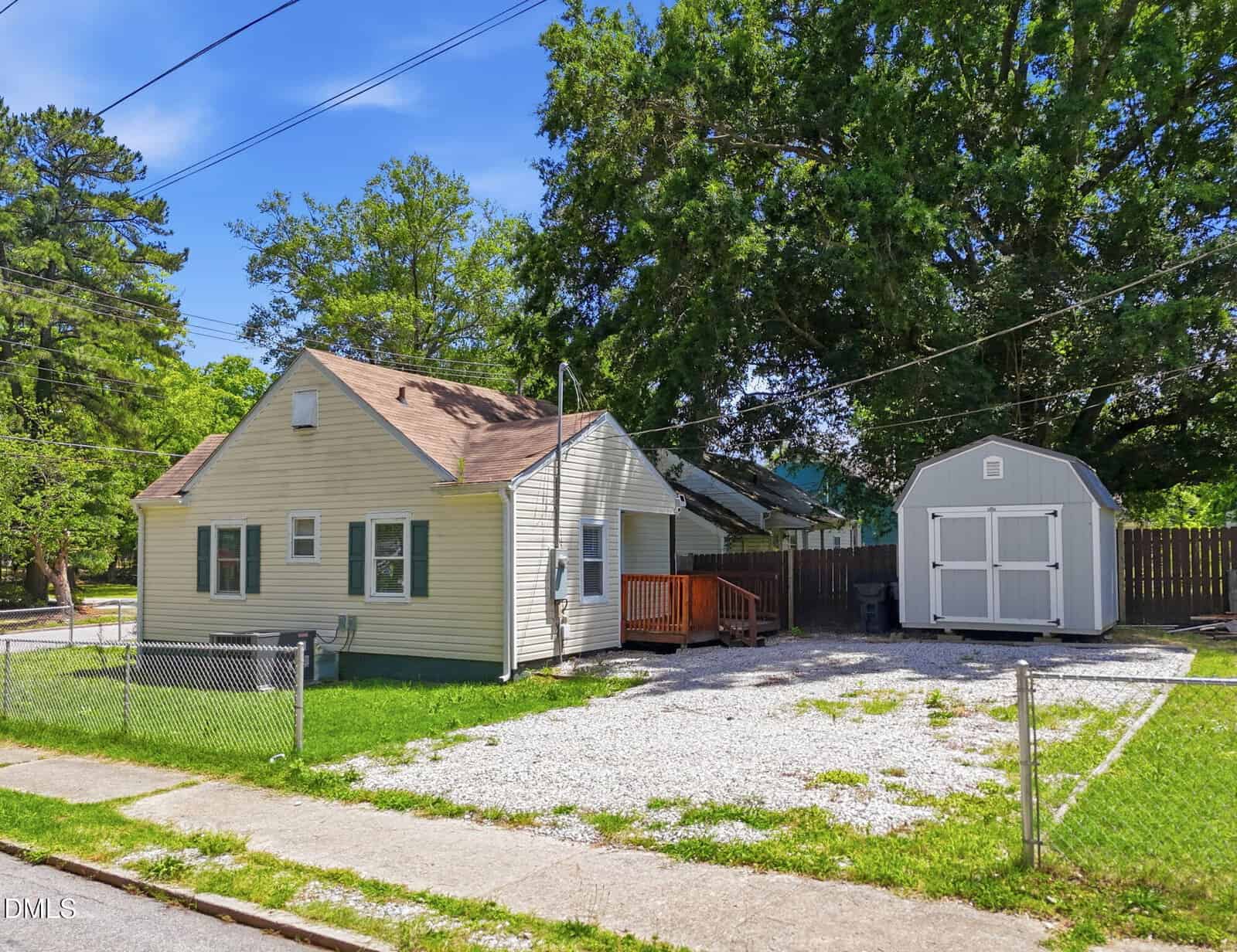 Single-story light beige house with a brown roof, small porch, and wheelchair ramp, beside a gravel driveway and a gray storage shed. Large trees and a chain-link fence border the yard. Bright, sunny day.