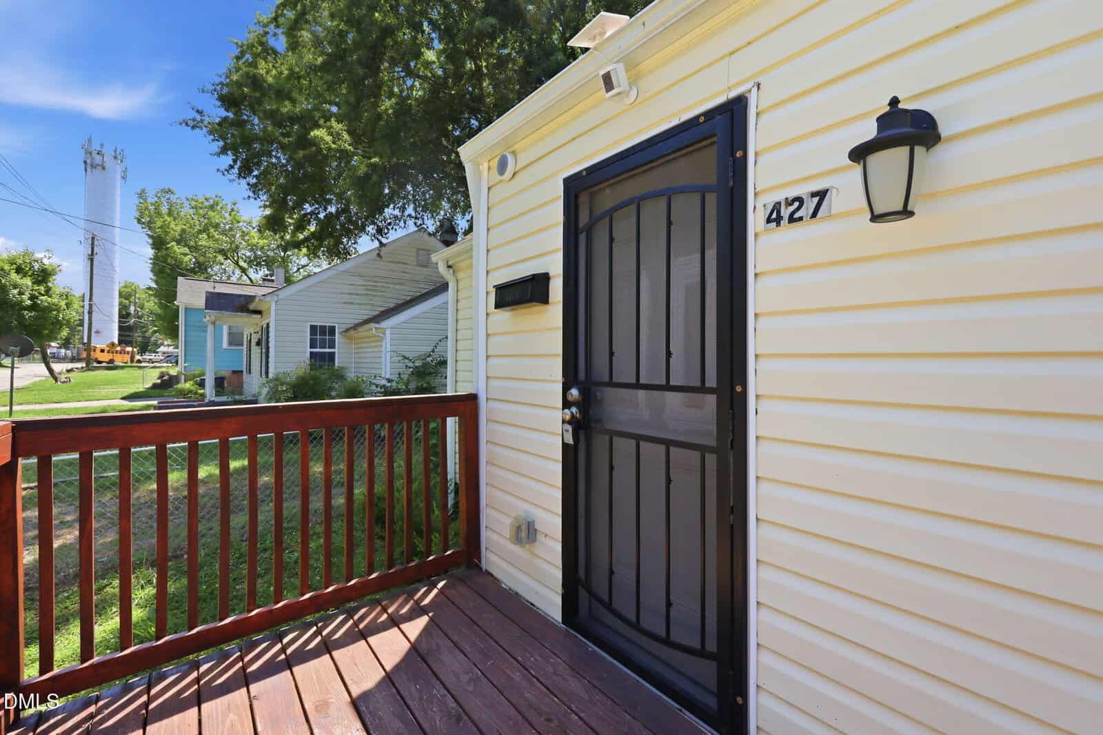 A close-up of a house entrance with a black security door, porch light, and house number 427. The house has cream siding, a red wooden deck, and a green yard with trees and neighboring houses visible.