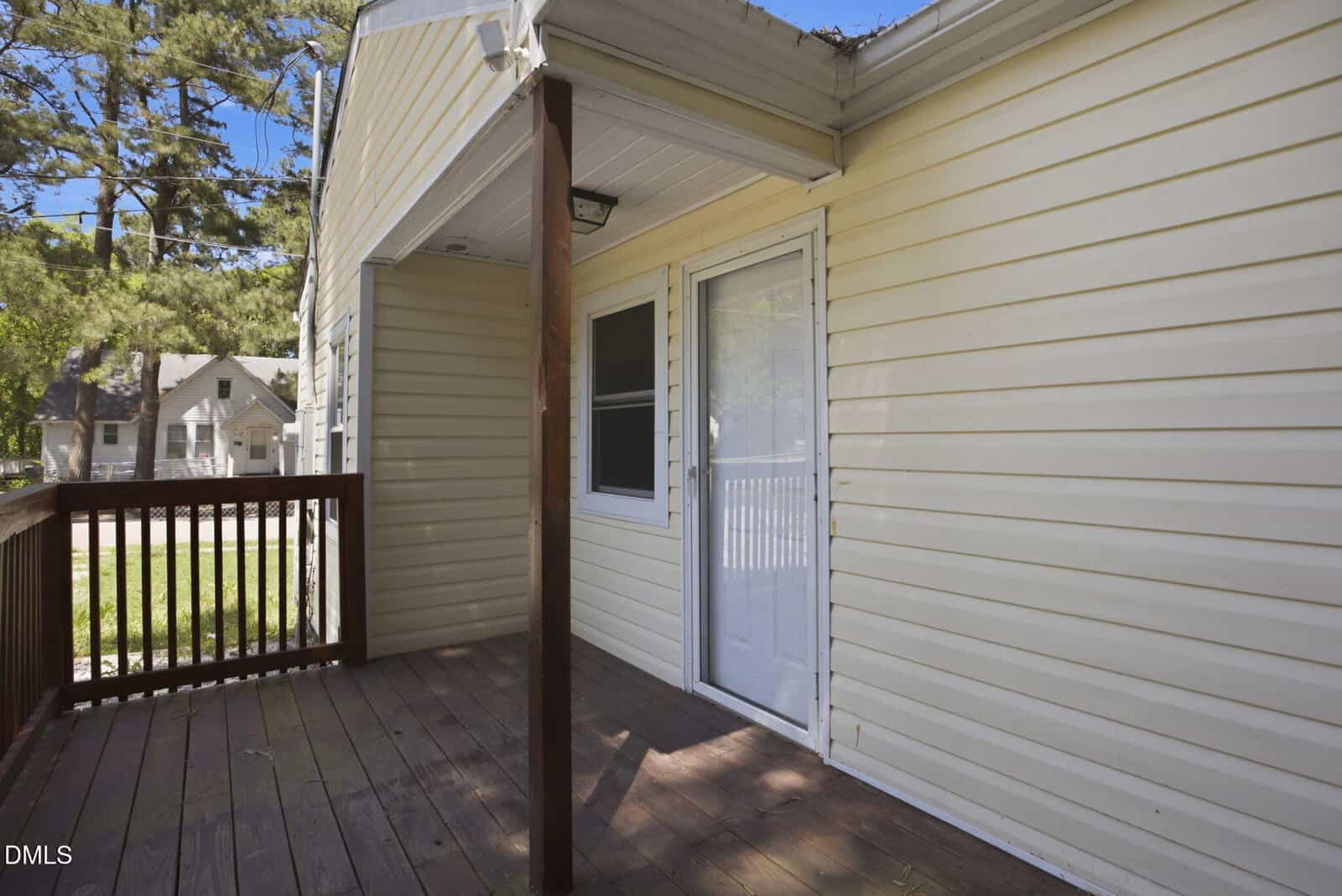 Small wooden deck with railing, attached to a beige vinyl-sided house. A white storm door with a window leads inside. The area is shaded by a small roof, with trees and neighboring homes visible in the background.