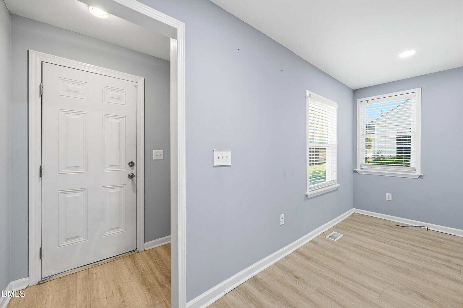 Entryway with a white door, light gray walls, two windows with blinds, light wood flooring, and recessed ceiling lights in a clean, empty room.
