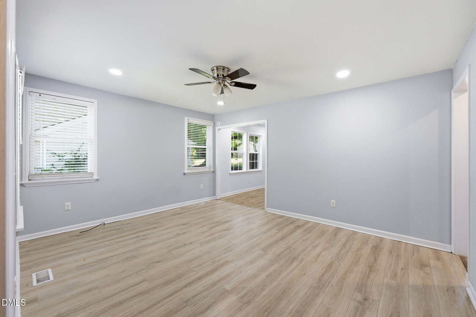 A bright, empty living room with light wood flooring, gray walls, white trim, recessed lights, a ceiling fan, and two windows. An open doorway leads to another room with similar flooring and windows.