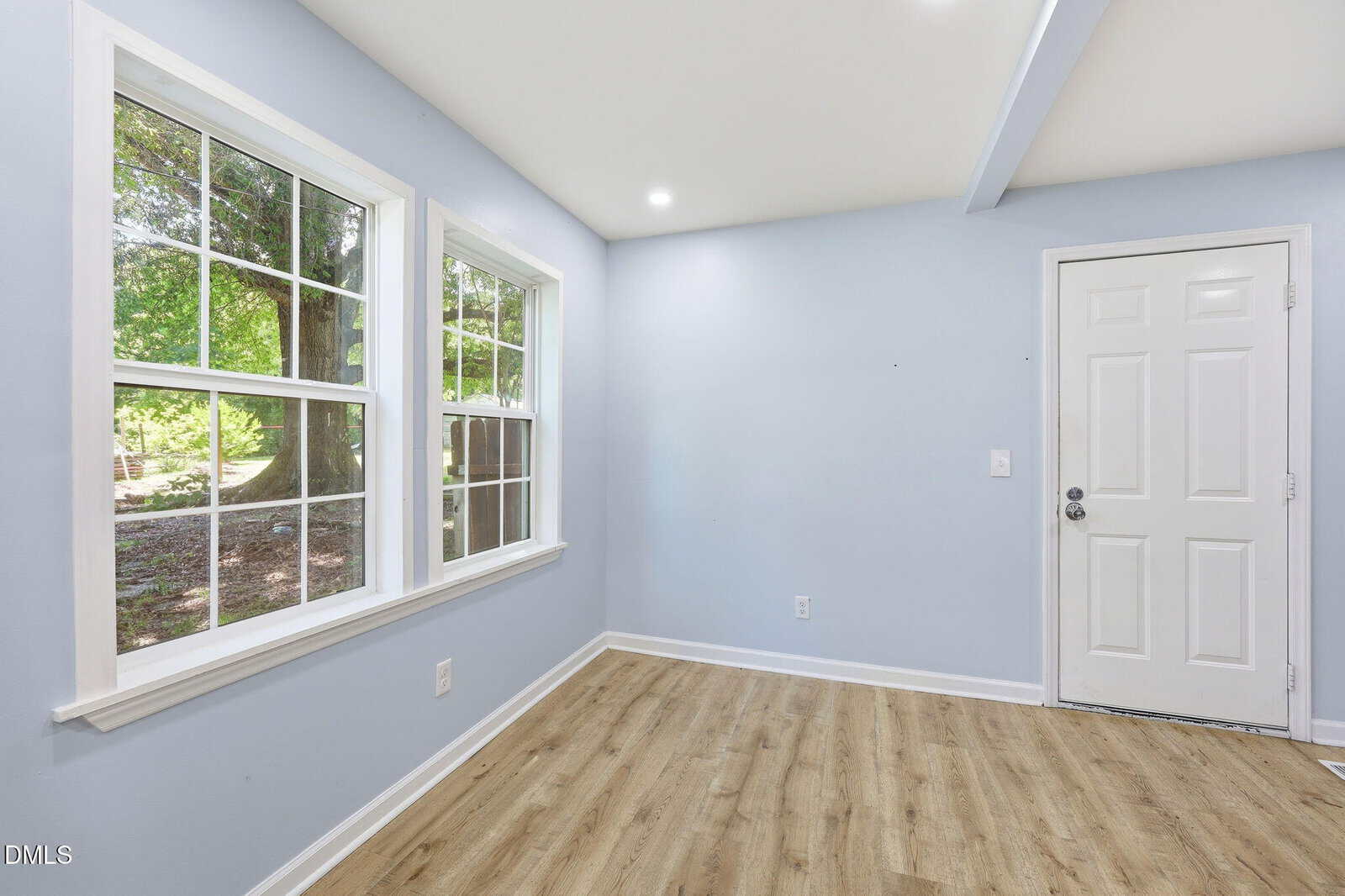 A bright, empty room with light blue walls, wood flooring, two large windows on the left, and a white door on the right. Sunlight shines through the windows, revealing a green outdoor view.