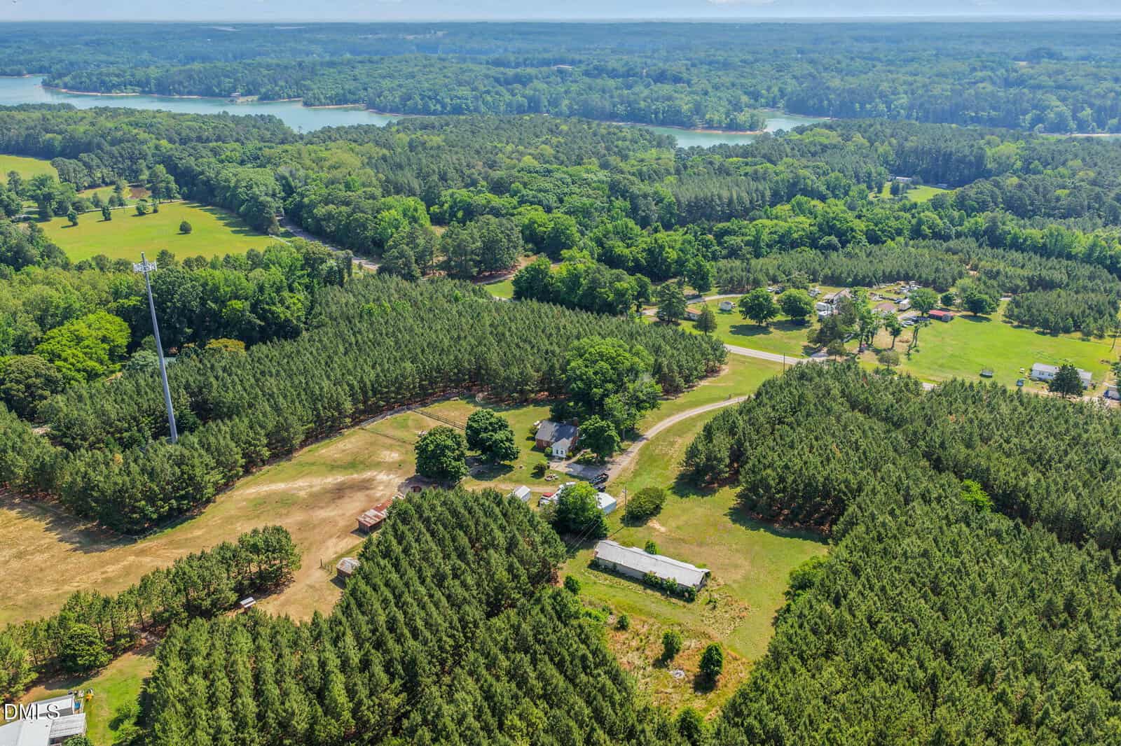 Aerial view of a rural landscape with dense pine forests, scattered houses, open fields, farm buildings, and a distant lake surrounded by trees under a clear sky.