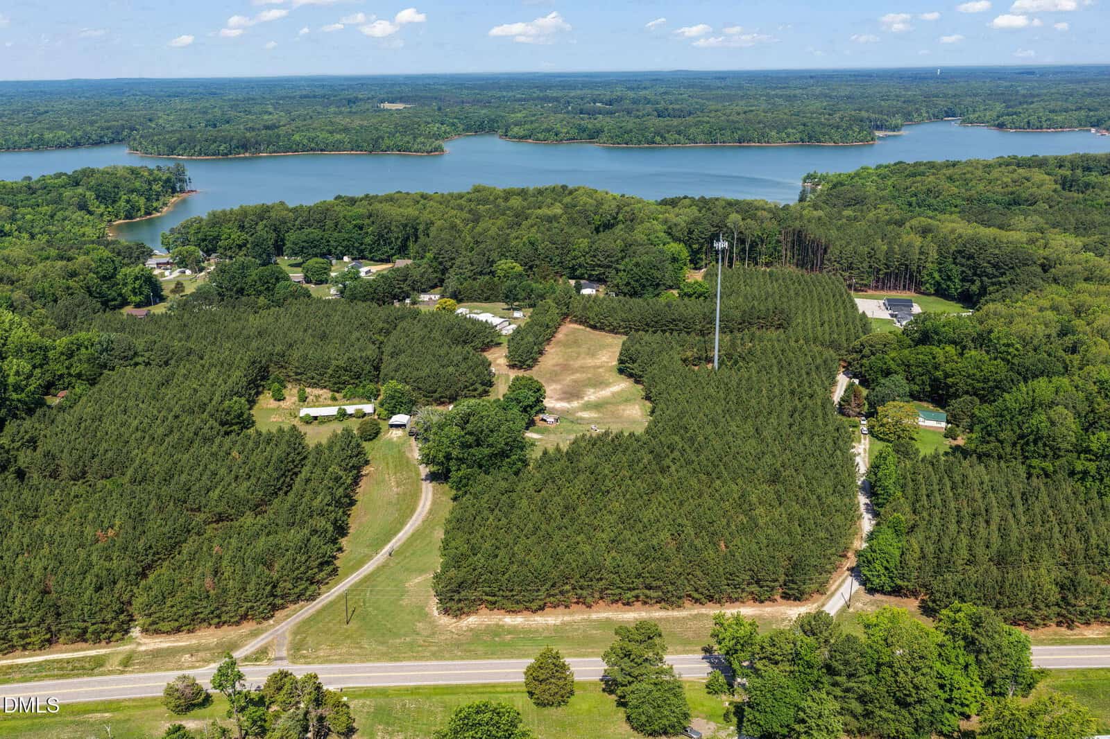 Aerial view of a rural landscape featuring dense pine forests, scattered homes, a tall cell tower, a winding road, and a large lake with a wooded shoreline in the background under a partly cloudy sky.