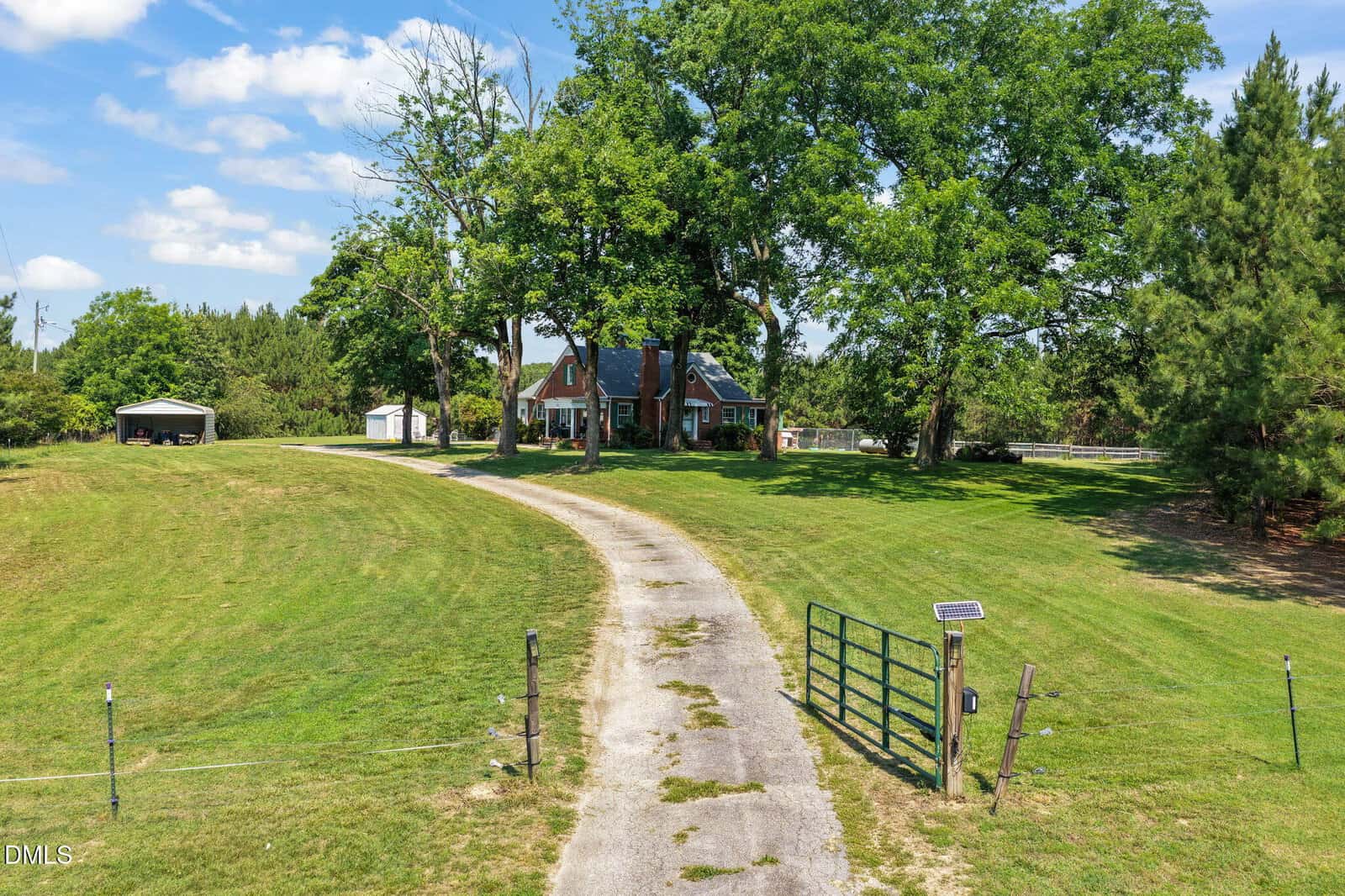 A long driveway leads to a brick house surrounded by trees and green grass, with a metal gate at the entrance and a small shed on the left side under a blue sky with clouds.