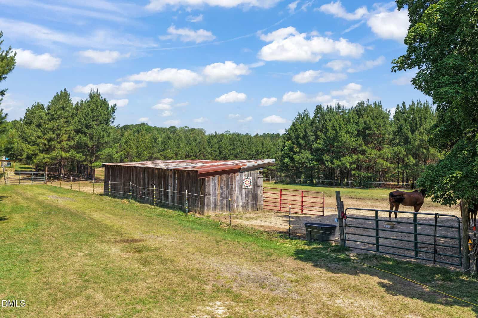 A brown horse stands in a fenced paddock beside a rustic wooden barn, surrounded by trees under a blue sky with scattered clouds.