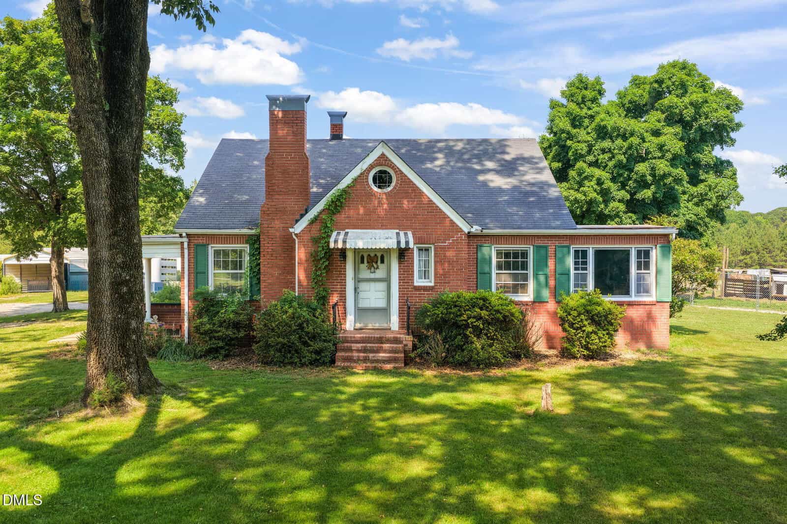 A red brick house with a gray roof, central chimney, and white front door with a small awning sits on green grass at 5225 Satterwhite Point Road in Henderson, surrounded by shrubs and tall trees under a partly cloudy sky.