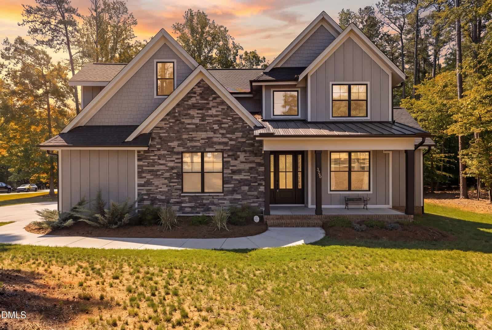 Nestled in Youngsville, 3790 Morning Song Court is a modern two-story house with gray siding, stone accents, black-framed windows, and a covered front porch, surrounded by trees and a grassy yard at sunset.