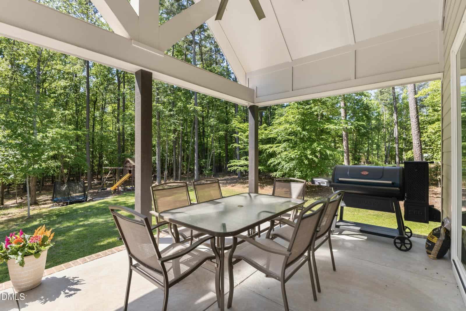 Covered patio with a glass table and six chairs, a grill to the right, potted plant on the left, and a view of a wooded backyard with a playset among the trees.
