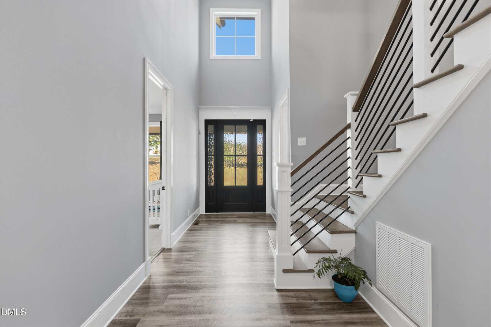 A modern entryway with light wood floors, gray walls, a black double front door with glass panels, a staircase with dark wood steps and black railings, and a potted plant at the base of the stairs.
