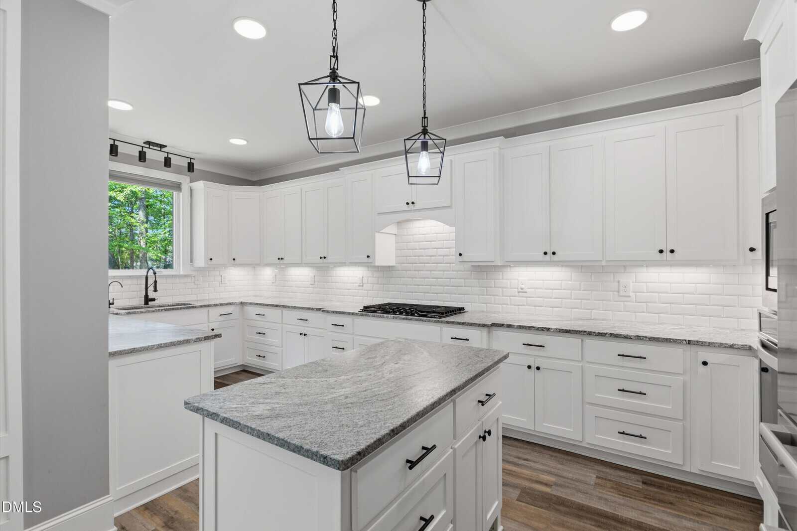 Bright modern kitchen with white cabinets, marble countertops, subway tile backsplash, island, stainless steel appliances, wood floor, and pendant lights. Large windows let in natural light and overlook greenery.