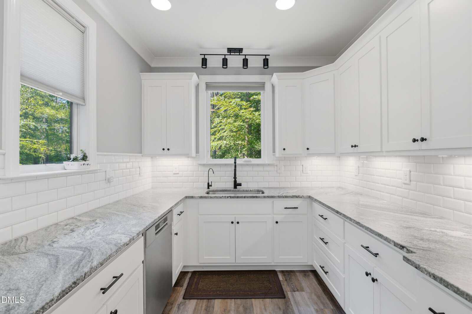 Bright, modern kitchen with white cabinets, marble countertops, white subway tile backsplash, stainless steel dishwasher, farmhouse sink under a large window, and wood-style flooring. Green trees are visible outside.