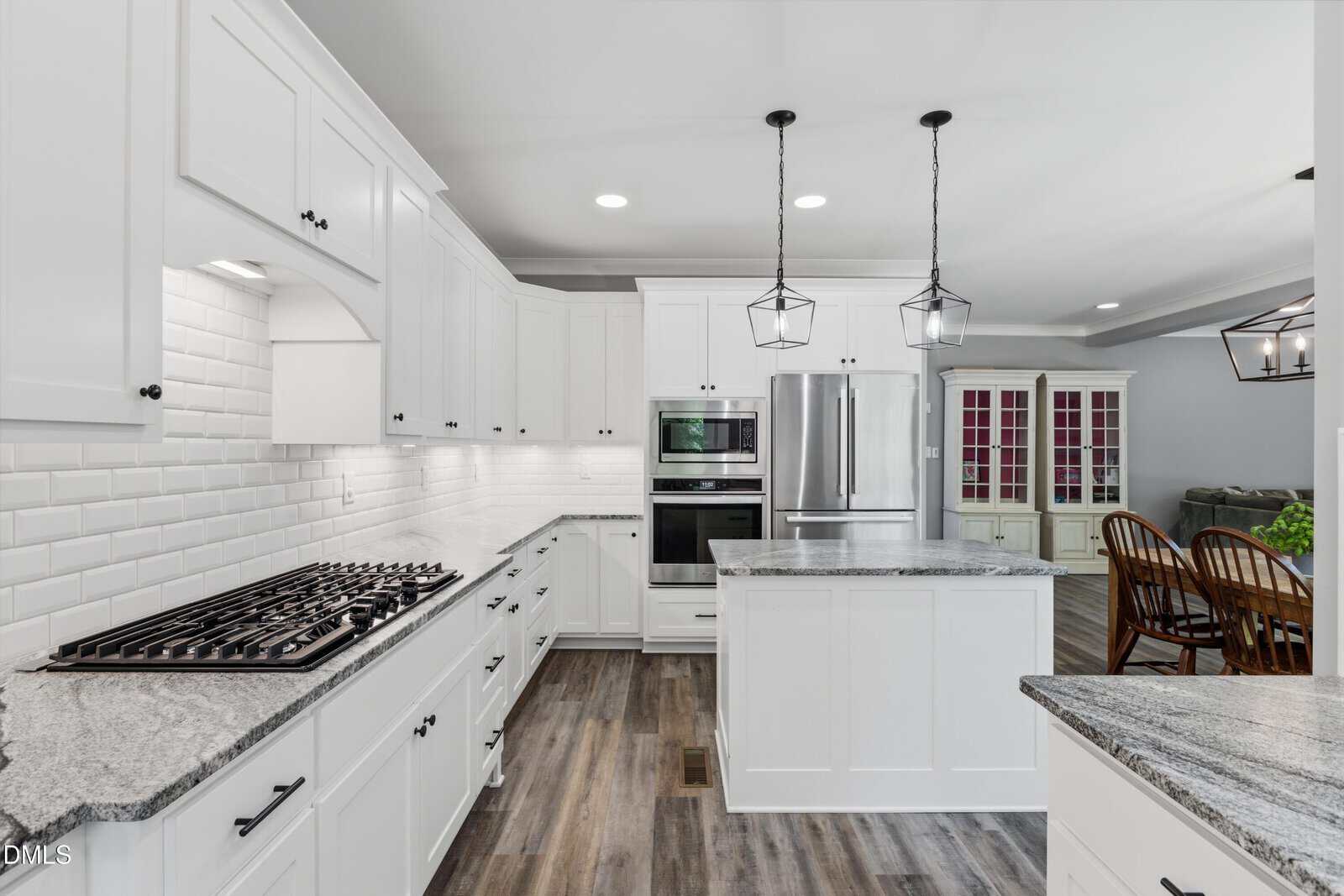 Modern white kitchen with stainless steel appliances, marble countertops, subway tile backsplash, a central island, pendant lights, and wood flooring. Dining area with wooden chairs and a hutch is visible in the background.