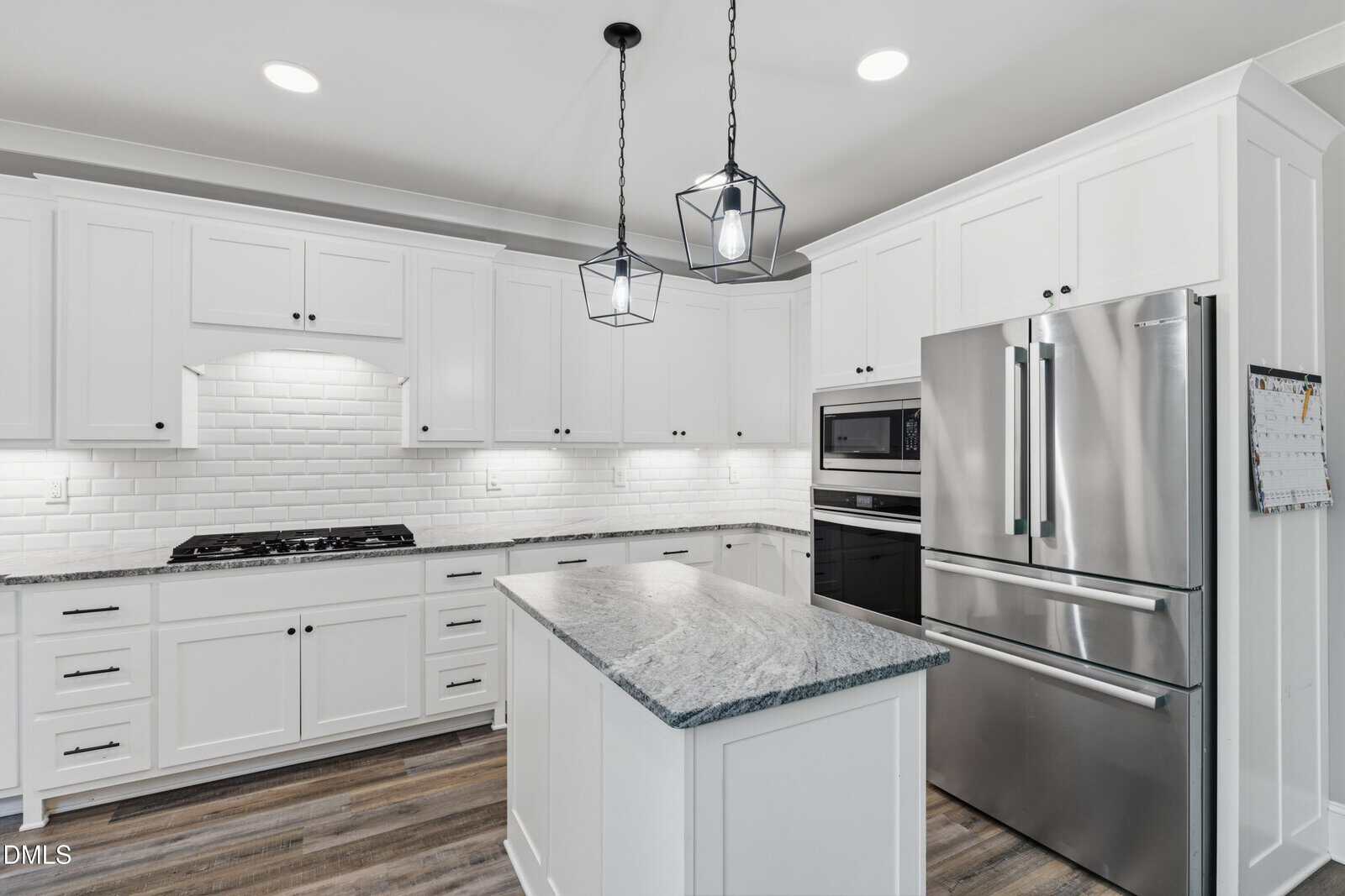 Modern kitchen with white cabinets, stainless steel appliances, marble island, and pendant lights. Subway tile backsplash and wood flooring complete the clean, bright look.