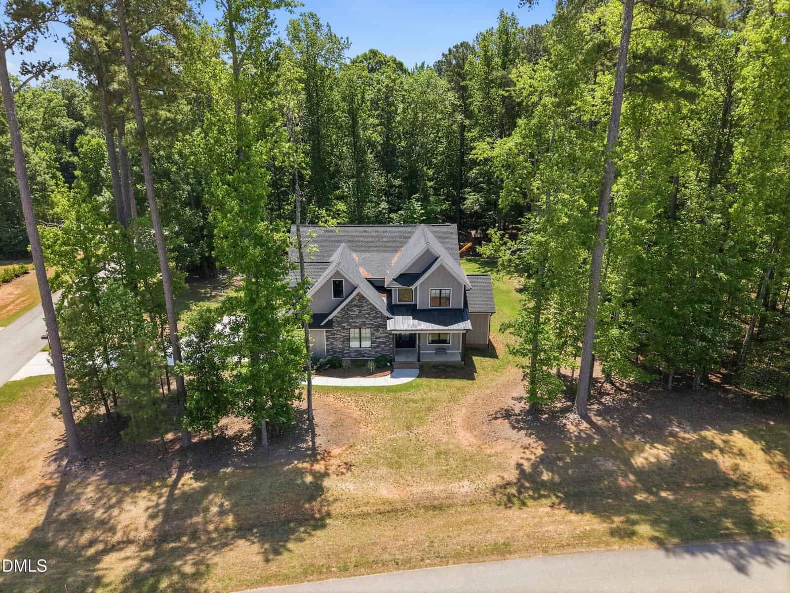 Two-story gray house with stone accents surrounded by tall trees on a spacious, grassy lot. The home features multiple roof peaks and sits near a quiet road under a bright, clear sky.