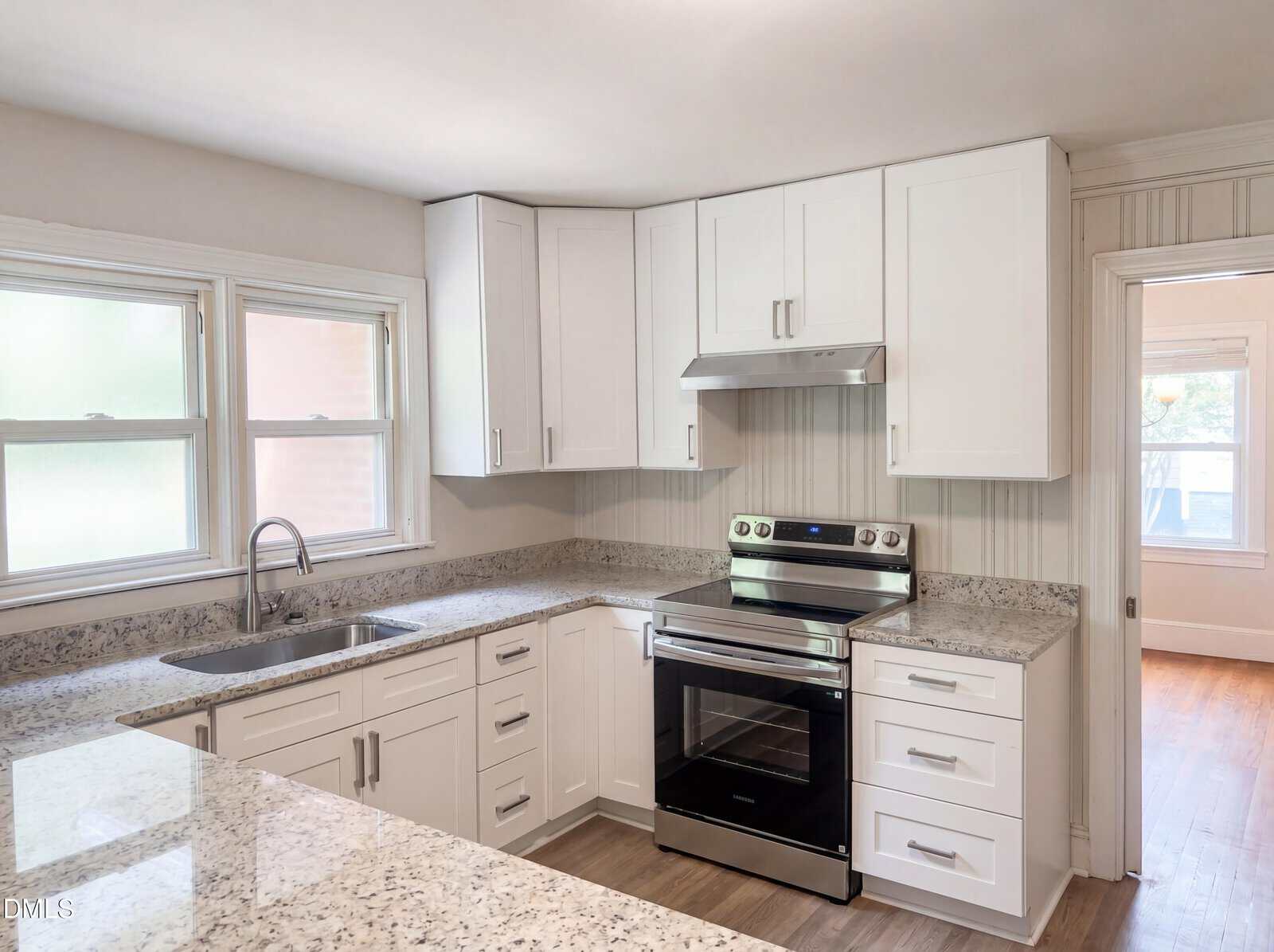 Modern kitchen with white cabinets, stainless steel oven and range hood, granite countertops, a double sink under two windows, and light wood flooring. An adjacent room with large window is visible through a doorway.