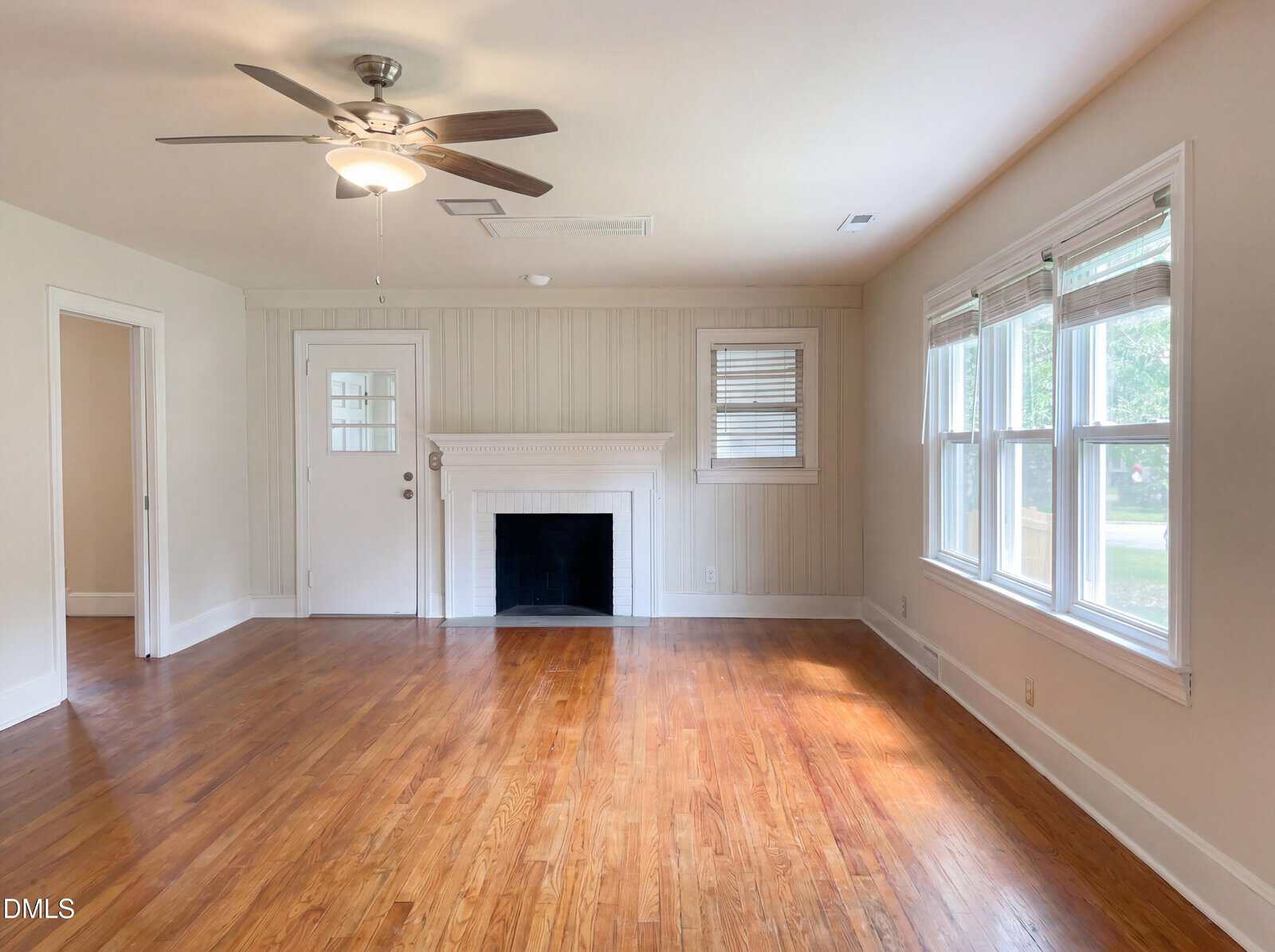 A bright, empty living room with hardwood floors, a ceiling fan, white walls, large windows, and a white fireplace with a mantel. Natural light streams in from the right side windows.