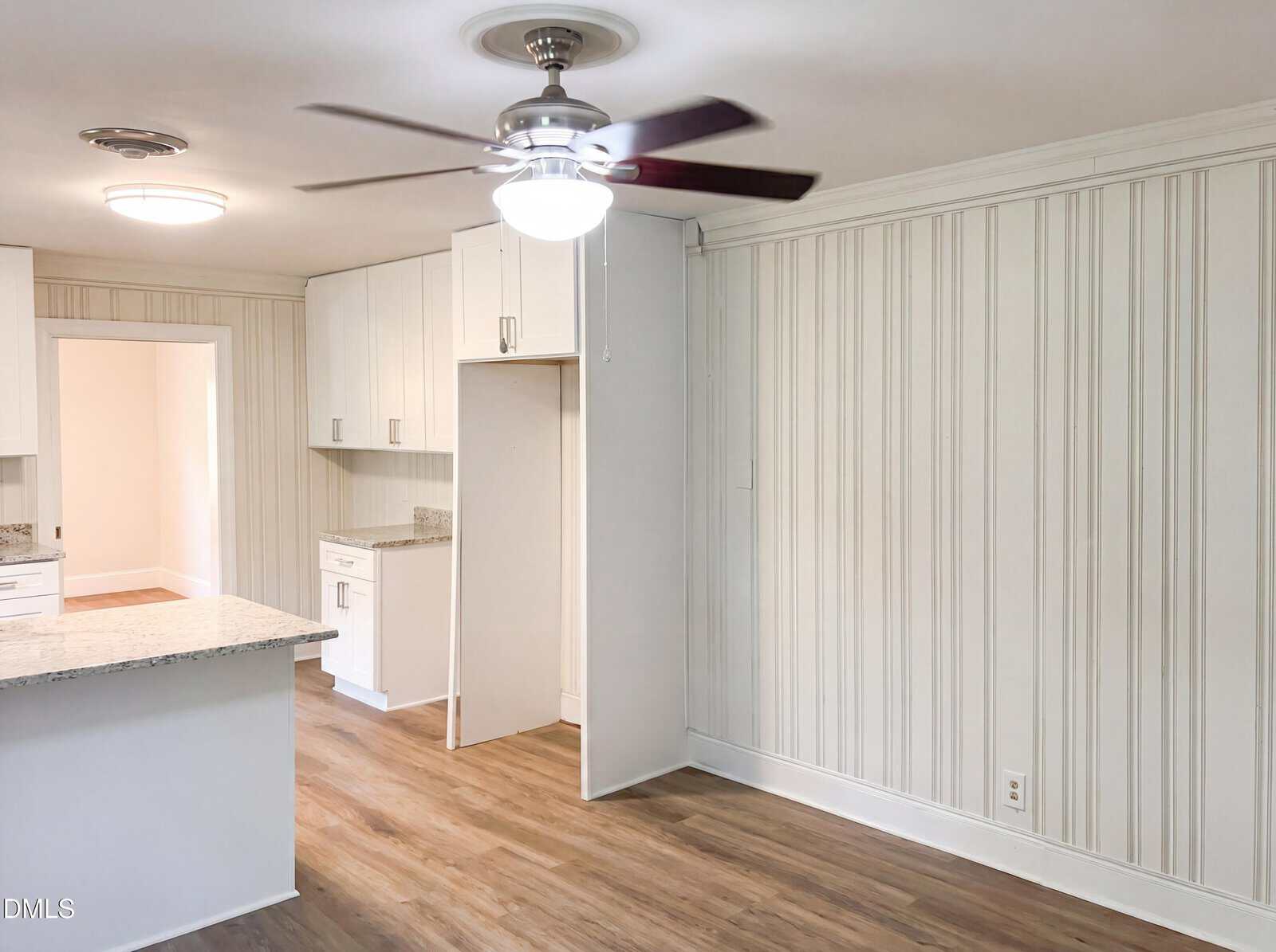 A bright, modern kitchen features white cabinets, wood flooring, granite countertops, vertical beadboard walls, and a ceiling fan with lights. An empty space is visible for an appliance, likely a refrigerator.