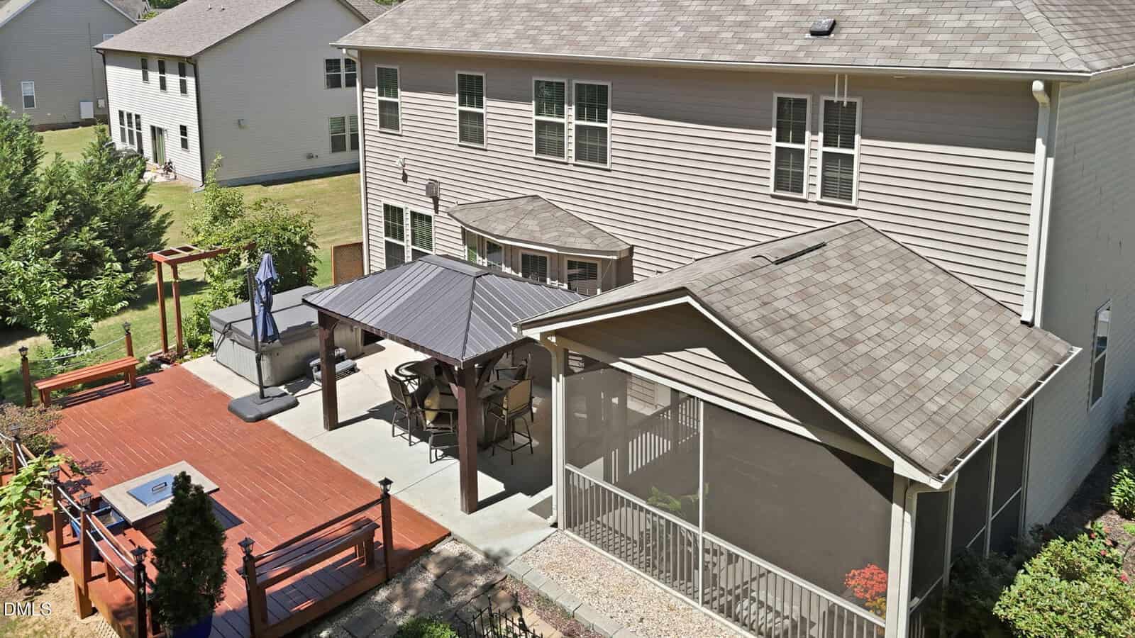 Backyard view showing a two-story house, a screened porch, a large patio with a gazebo, hot tub, dining set, and a wooden deck bordered by plants, all surrounded by grassy lawns and neighboring homes.